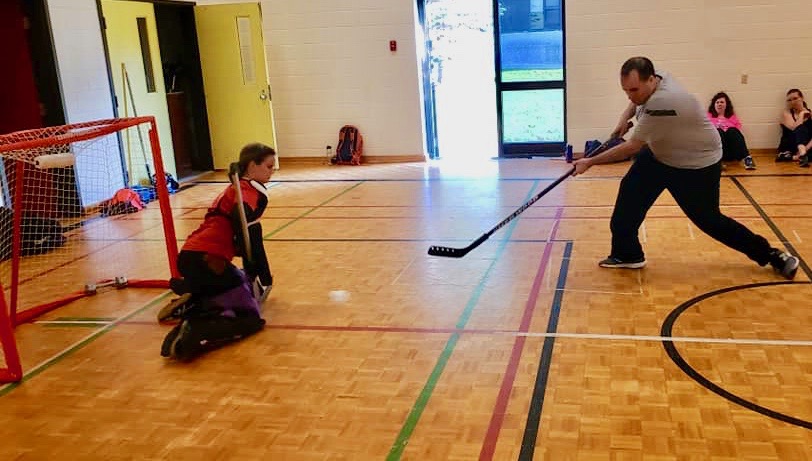 A goalie is positioned in front of a hockey net as a mother shoots the ball in that direction 