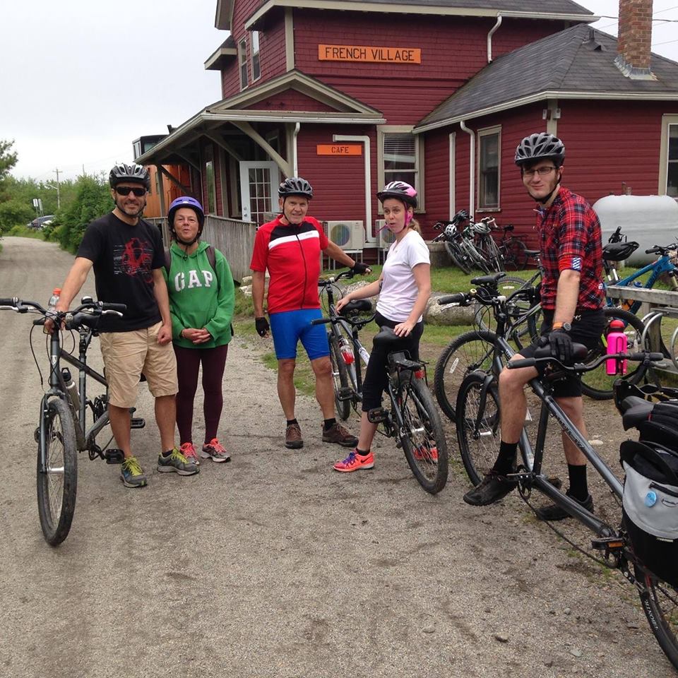 A group of 5 people stand with tandem bikes on a trail in front of a cafe