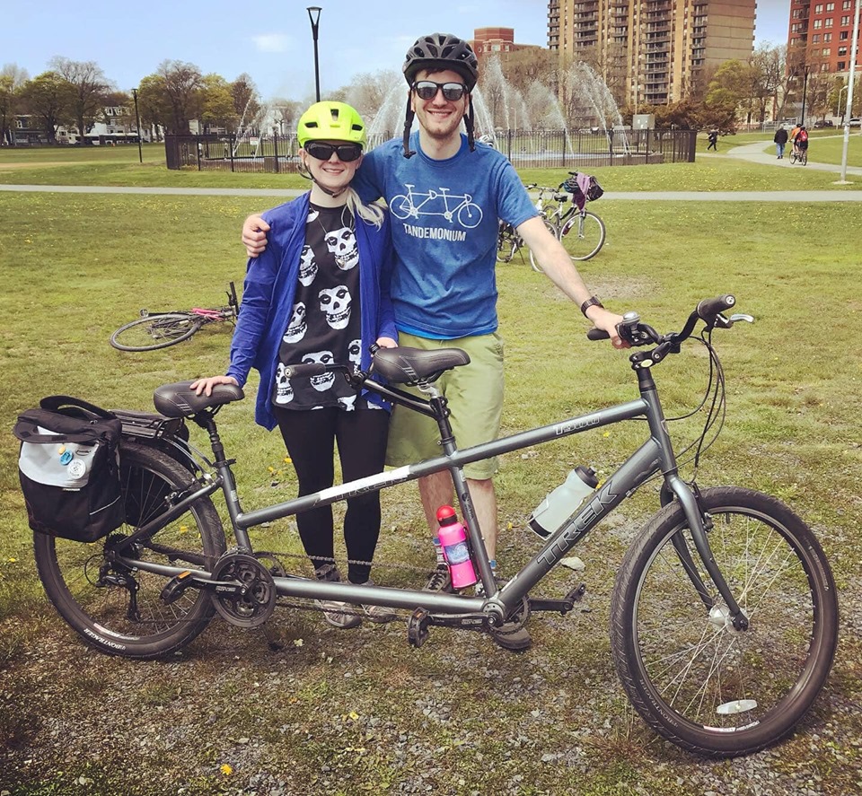 2 people with tandem bike. They're in a park and smiling.