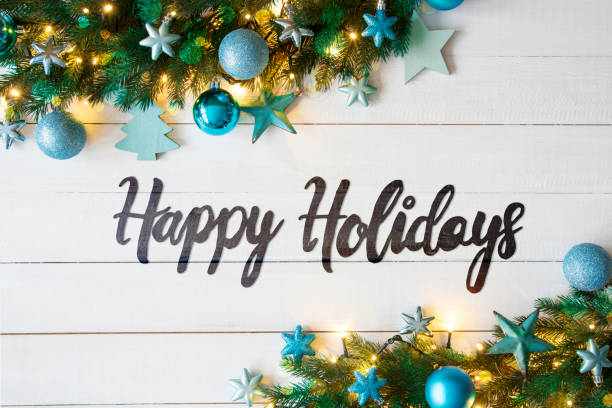 Happy holidays in grey-black handwriting style text in front of whitewashed wood and bordered by evergreen leaves, blue-themed holiday ornaments (like stars, trees, and snowflakes) and white lights
