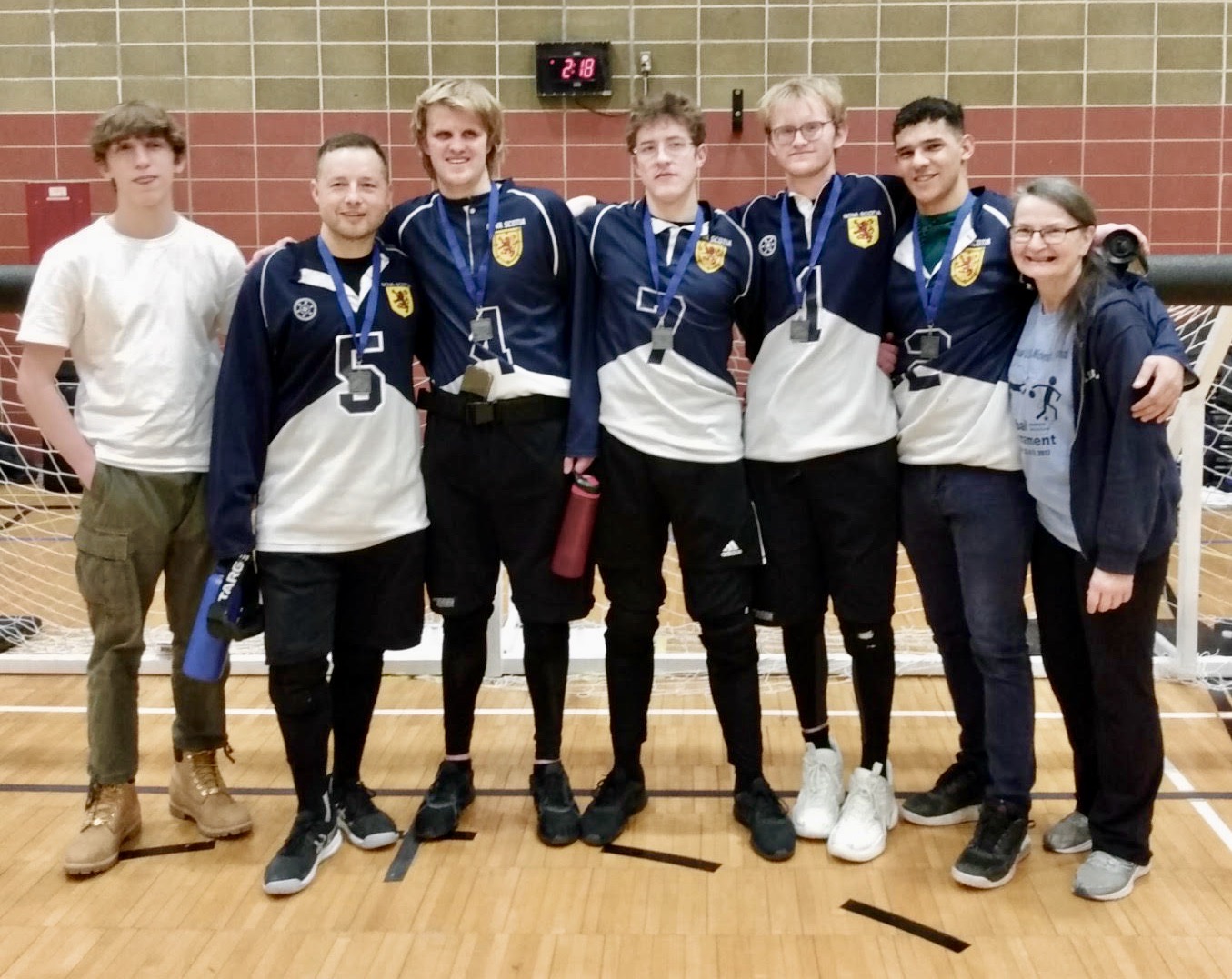 7 people in front of a goalball net in a gymnasium, wearing medals and smiling