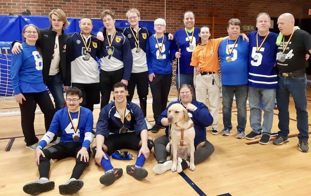 14 people wearing blue jerseys and medals around their next stand and sit in a group with arms around each others shoulders. A guide dog sits on the gymnasium floor.