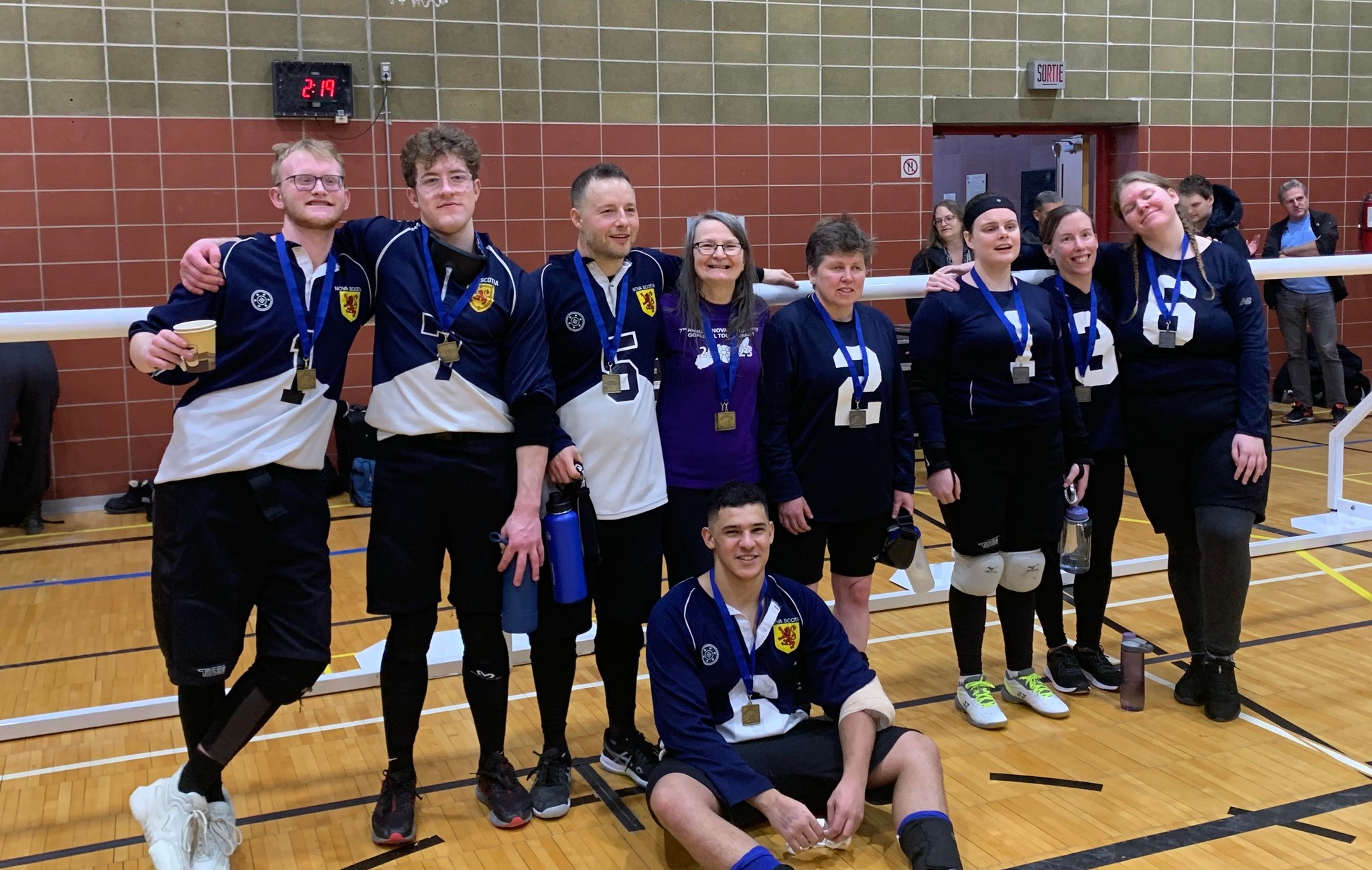 Nine people wearing medals around their next and smiling. Eight of them stand against a long goalball net and one sits on the floor in front of them.