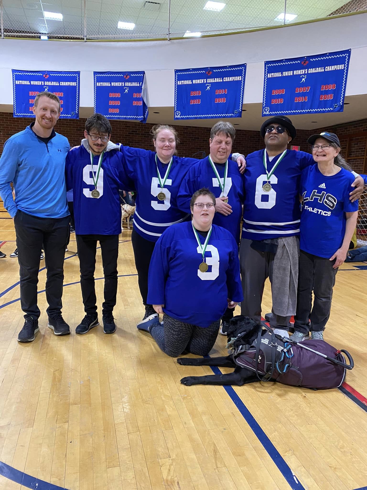 A group of people wearing gold medals in a gymnasium, a dark coloured guide dog stands in front.
