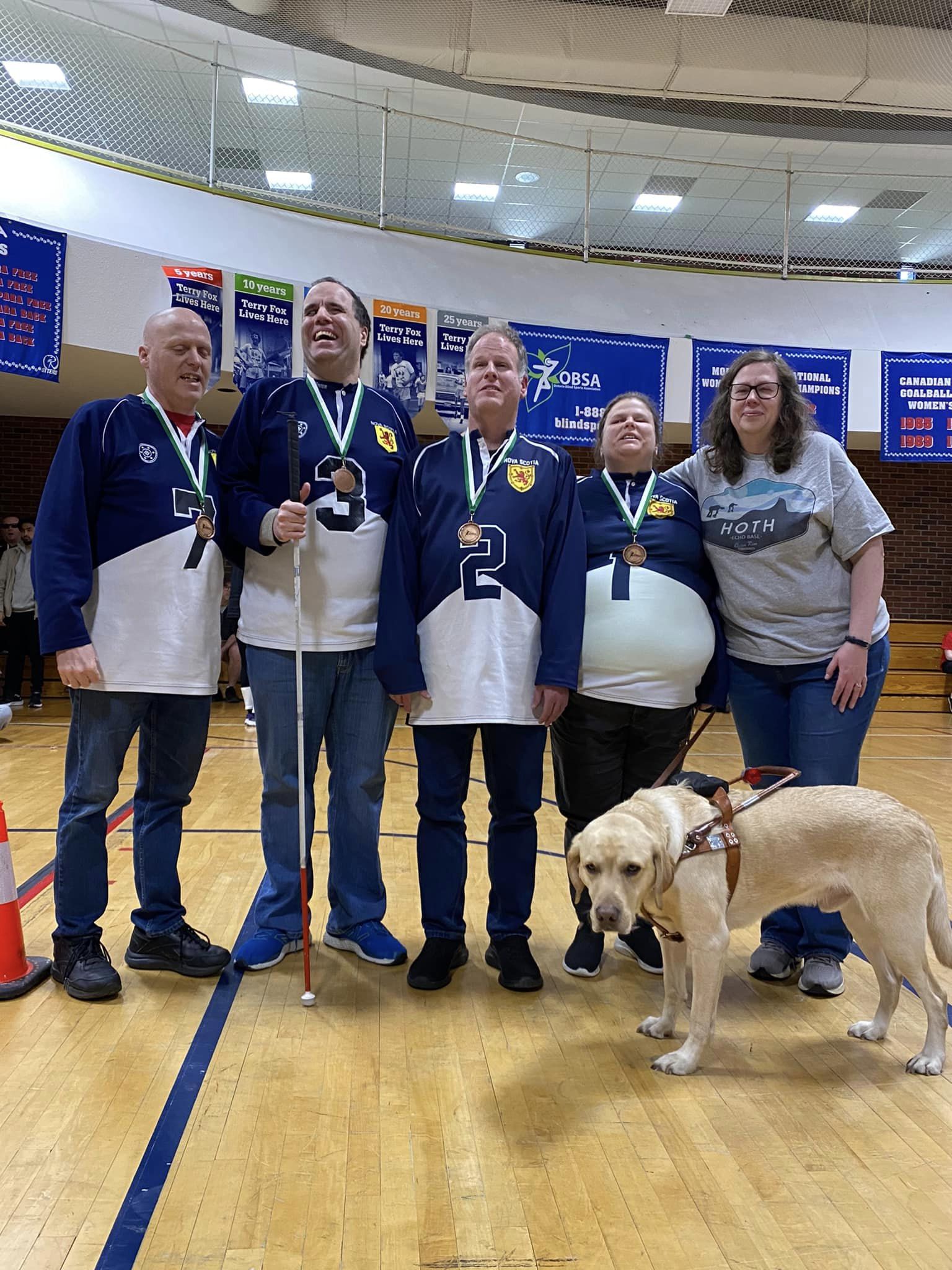 A group of people standing in a gymnasium wearing bronze medals, a golden coloured guide dog is in front