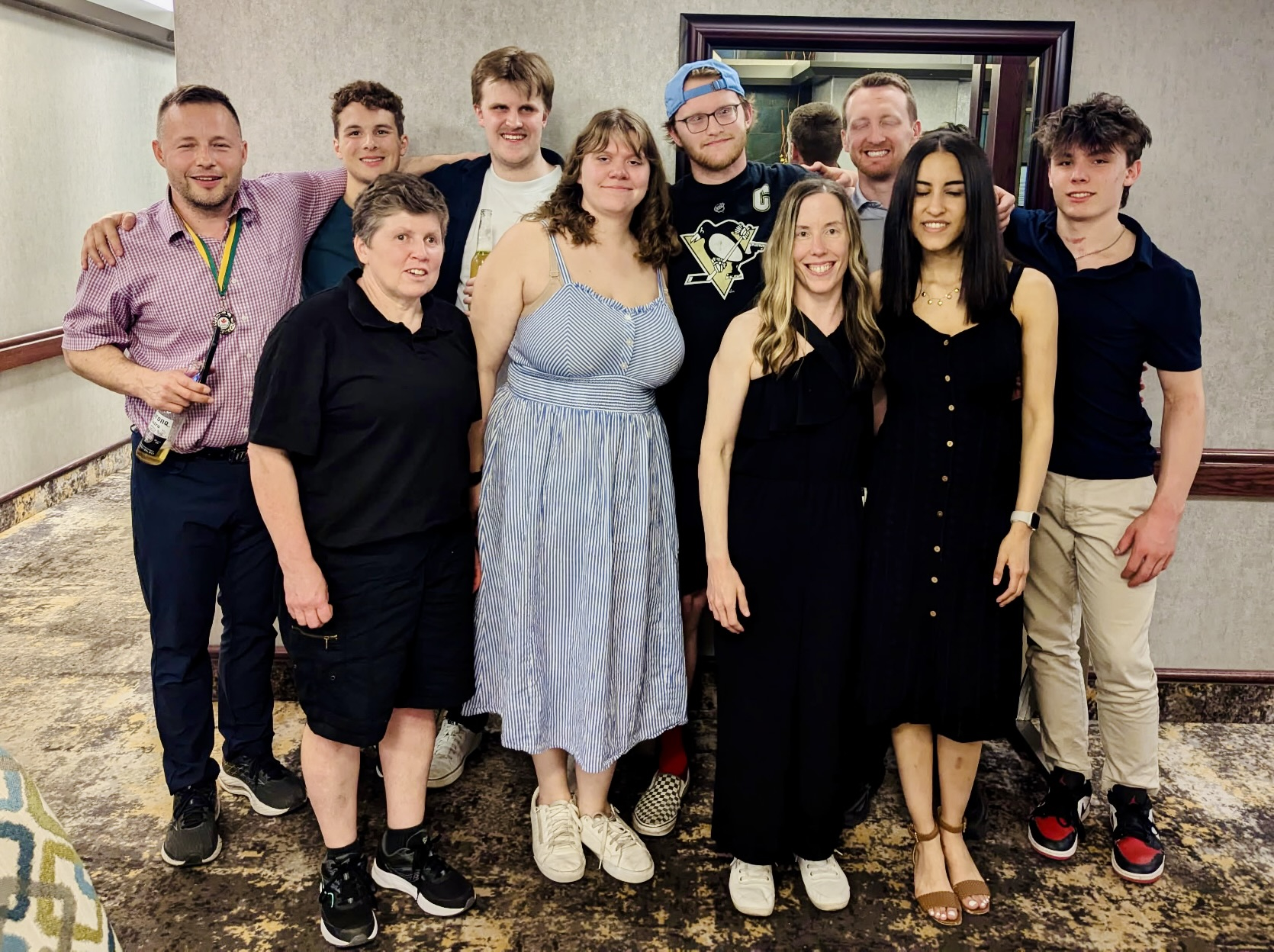 A group of ten people stand shoulder to shoulder in 2 rows, smiling and many with arms around one another. They stand against a light background with a mirror on dark patterned carpeting.