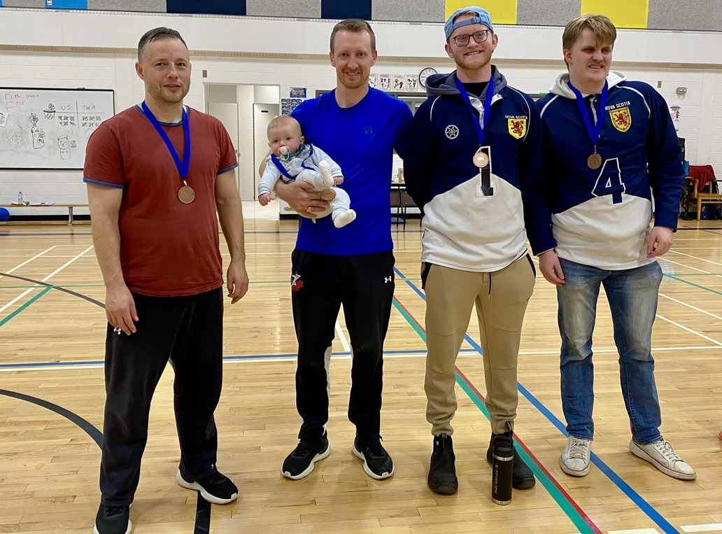 A group of 4 men with bronze medals around their necks stand smiling in a gymnasium. Two are wearing blue & white sports jerseys and the man second from the left is holding a small baby in his arms.