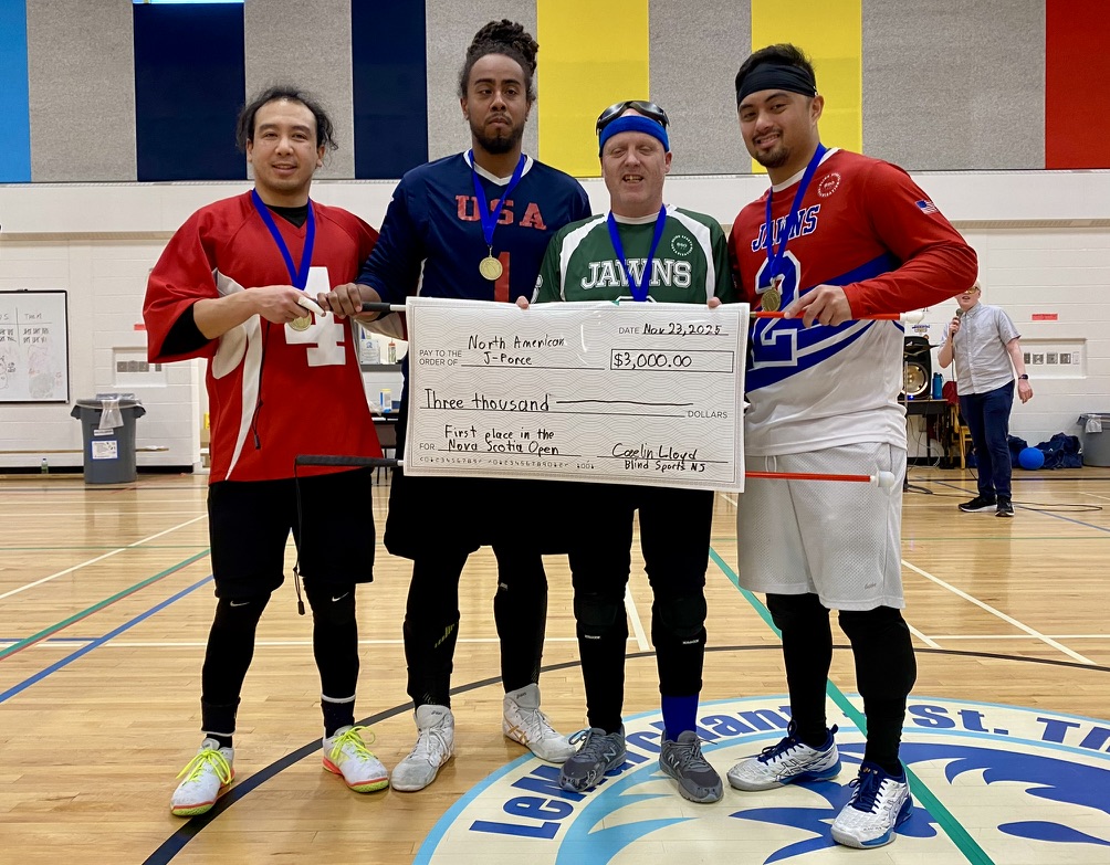 In a gymnasium four men stand shoulder to shoulder holding an oversized cheque for $3,000. They are smiling and wearing gold medals around their necks.