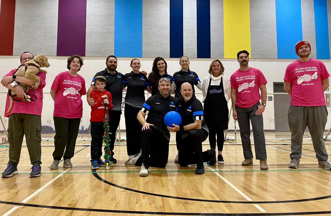 A group of 12 people in a gymnasium, some wearing matching pink t shirts, some collared uniform shirts. Many are smiling and one person to the left is holding a large golden puppy in their arms.