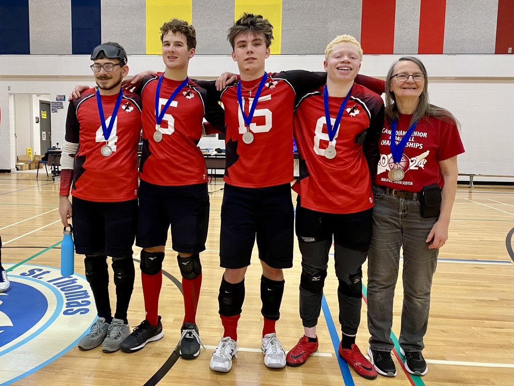 Four young men and one woman stand with arms around each other in a gymnasium. The four men are wearing red sports jerseys. All wear silver medals around their necks and are smiling.
